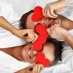 Paar in weißen Bademänteln hält rote Herzformen vor ihre Augen und lächelt vor Paarmassage.