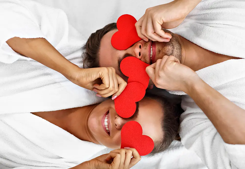Paar in weißen Bademänteln hält rote Herzformen vor ihre Augen und lächelt vor Paarmassage.
